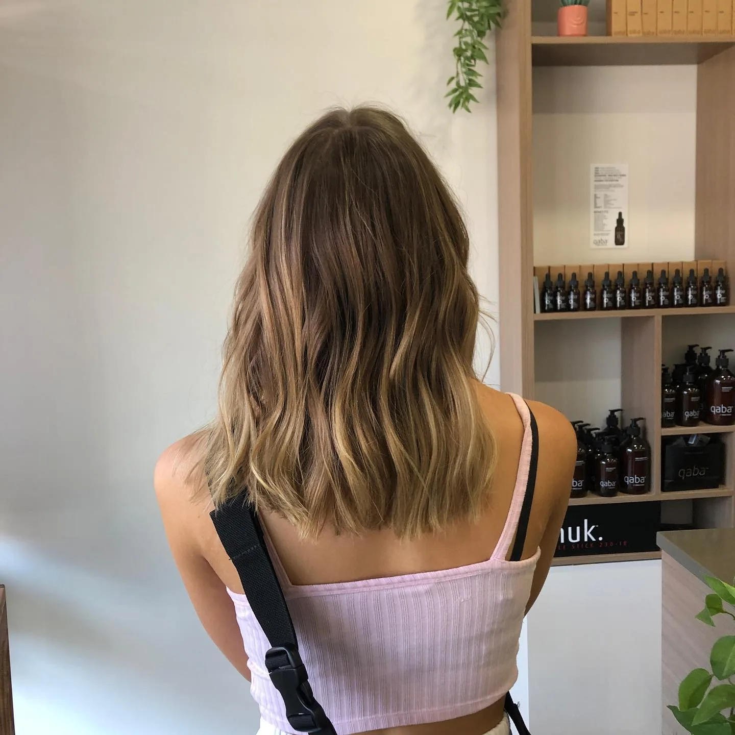 Retail shelf of amber qaba dropper and pump bottles behind a client with blonde balayage